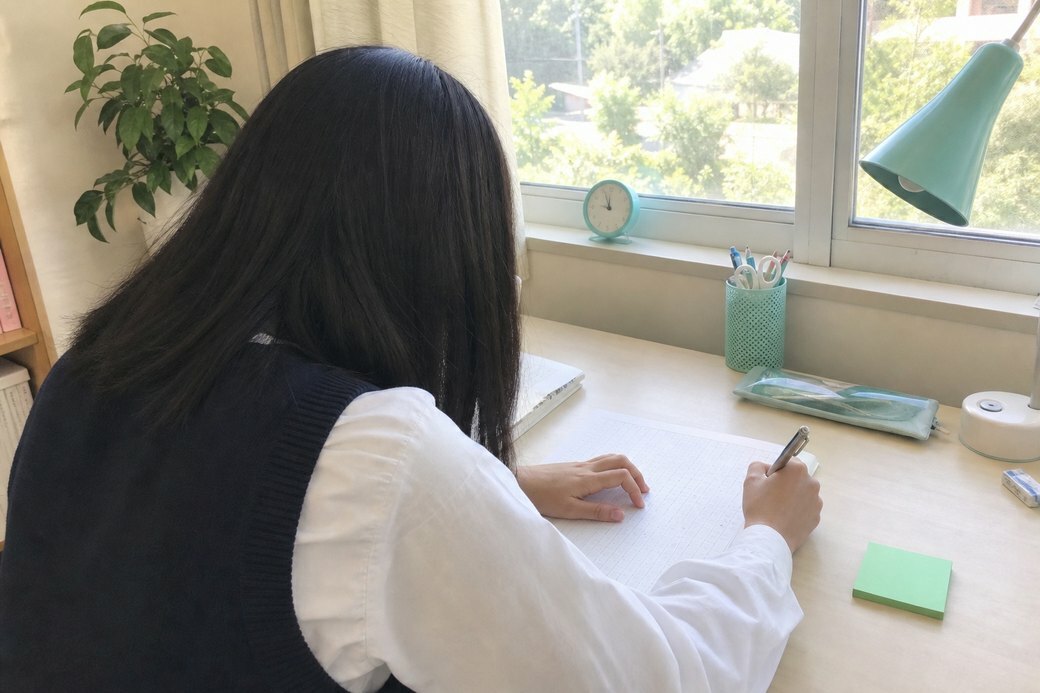 明るい窓辺の自習席で小論文に取り組む高校生の後ろ姿