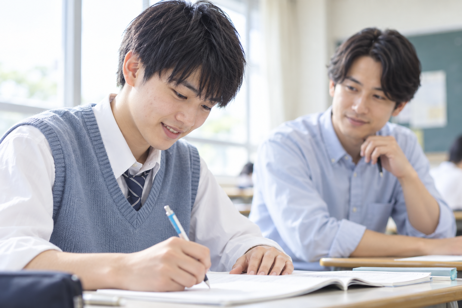 学習を進める男子高校生と、奥から見守る講師のイメージ